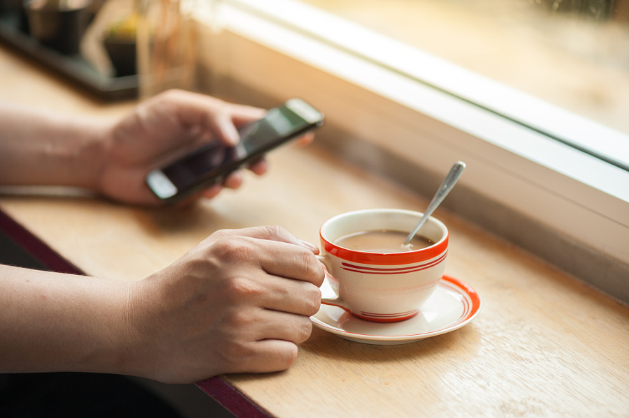 Male hand holding a cup of coffee on wood bar with another hand using smartphone in blurry background with morning scene
