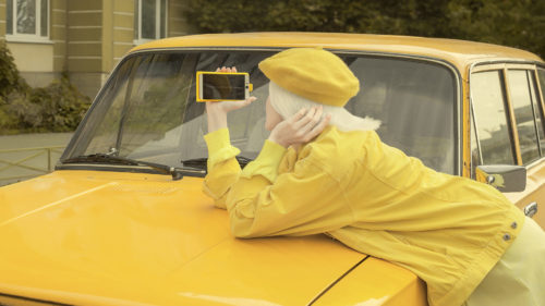 A woman wearing a yellow beret and yellow clothing leans on the hood of a yellow car, facing away from the camera