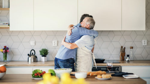An adult hipster son and senior father indoors at home, hugging when cooking