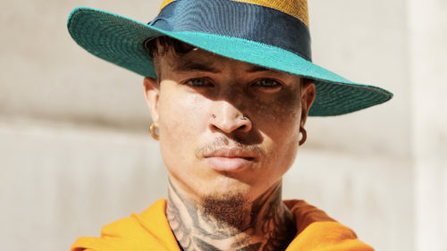 Man in a yellow hoodie with a neck tattoo and a hat looking at the camera