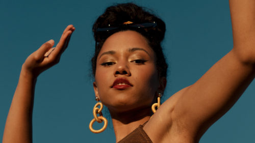 Back woman with her hair up wearing gold earrings dancing against a blue sky