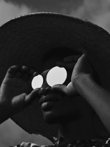 A person in a straw hat holds reflective discs over their eyes, captured in black-and-white with striking contrast and moody lighting.