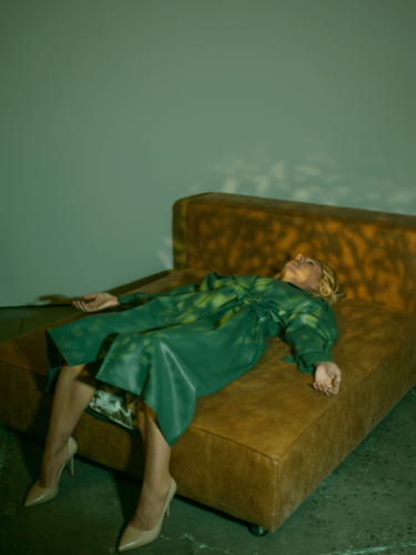 A woman in a green dress reclines on a tan leather sofa under dappled light, demonstrating color blocking in photography with bold wardrobe and muted surroundings.