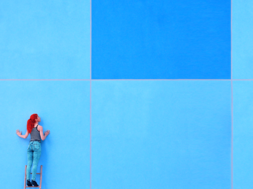 A woman with red hair stands on a ladder against a large blue tiled wall, emphasizing color blocking in photography with vivid, complementary colors.