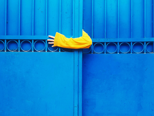 A hand in a yellow sleeve reaches through a bright blue gate, highlighting minimalist color photography with bold, contrasting tones.