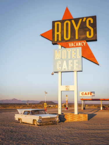 A classic Route 66 roadside scene featuring Roy's Motel Café sign and a vintage car, photographed with strong vintage aesthetics that evoke mid-century Americana.