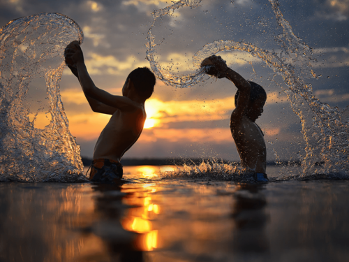 Two children silhouetted against a golden sunset, splashing water in dramatic arcs.