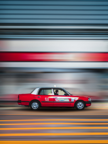 A red taxi in Hong Kong sharply focused against a streaked background, showcasing the panning technique to convey speed.