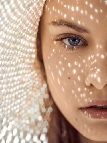 Close-up portrait of a woman with blue eyes wearing a hat, dotted light patterns on her face, showcasing midday sunlight photography.