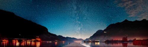A breathtaking nightscape by Sirun Tang showing the Milky Way above a still lake, framed by mountains and dotted with warm lights along the shore.