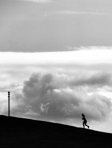 Black and white photo by Nicole Grenier of a lone runner silhouetted on a hill against dramatic clouds, evoking solitude and resilience.
