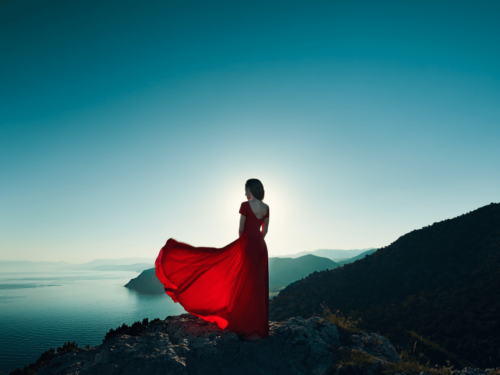 Woman in a flowing red dress standing on a mountain cliff with a twilight view over the sea.