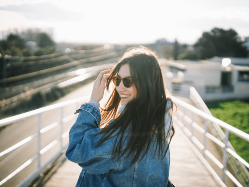 Smiling woman in sunglasses and a denim jacket walking outdoors on a bright day, captured with harsh lighting photography.