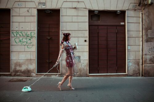 Street photography by Olena Leliuk showing a woman in a transparent dress walking down a city street, pulling an iron on a cord like a pet.