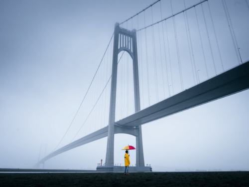 A person in a yellow raincoat stands under a suspension bridge holding a colorful umbrella on a foggy morning, capturing the mood of rain, fog, and overcast skies.