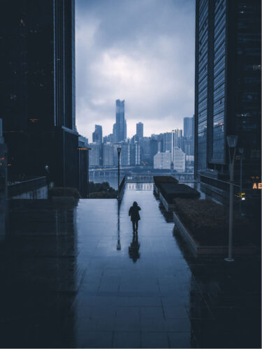 A lone figure walks along a reflective, rain-soaked urban path framed by tall skyscrapers under heavy clouds, capturing the mood of rain, fog, and overcast skies.