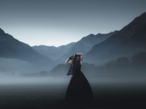 A woman in a flowing black gown stands in a misty valley surrounded by mountains at dusk, capturing the mood of rain, fog, and overcast skies.