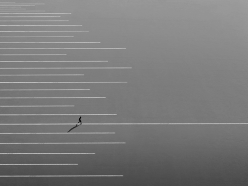 A lone figure walks across an empty parking lot with long diagonal lines creating a minimalist scene, composing for mood through isolation and shadow.