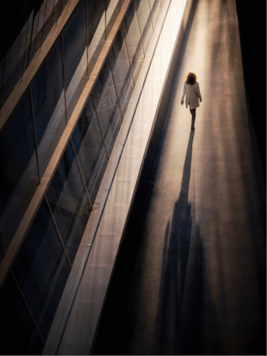 A person walking along a sunlit urban path with a long shadow, highlighting dramatic lighting and geometric composition.
