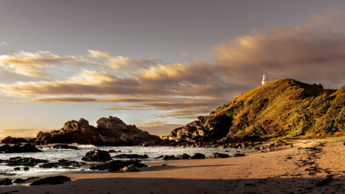 Coastal landscape photo by Graeme Ian Hall capturing a lighthouse on a sunlit hill above a rocky beach during golden hour with dramatic clouds.