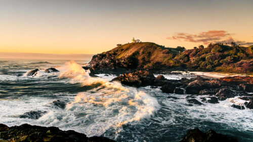 Seascape photo by Graeme Ian Hall showing waves crashing against rocky shores beneath a golden sunrise, with a lighthouse on a hill in the distance.