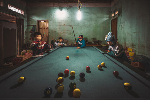 Documentary photo by Marco Tagliarino of children playing billiards in a dimly lit room, capturing a candid moment of everyday life.
