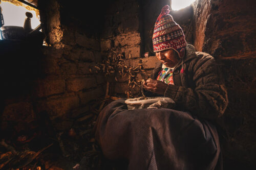 Candid portrait by Marco Tagliarino of an elderly woman in traditional clothing working by a window inside a dim, rustic room.