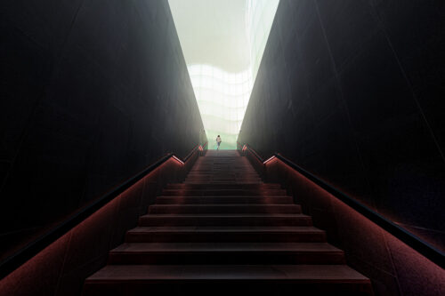 Architectural photo by Marco Tagliarino of a lone figure ascending a dramatic staircase framed by tall dark walls and glowing red handrails.”