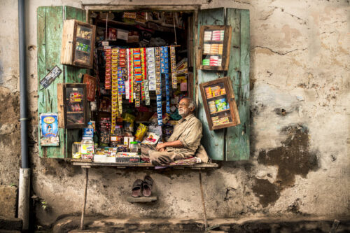 Street portrait by Marco Tagliarino of a shopkeeper sitting inside a small, colorful market stall framed by weathered green shutters.