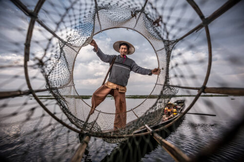 Travel portrait by Marco Tagliarino of a fisherman balancing on a wooden boat, framed through a large circular fishing net on a calm lake.
