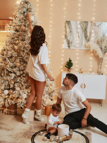 A young family decorating a snowy Christmas tree in a cozy, warmly lit living room while their toddler plays with ornaments, creating a scene of holiday storytelling.