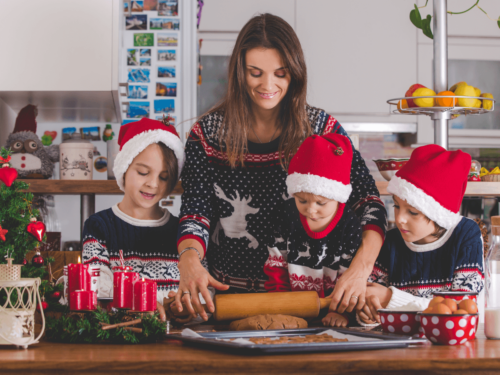 A mother and three children in matching Christmas sweaters and Santa hats rolling cookie dough together in a festive kitchen, capturing a warm moment of holiday storytelling.