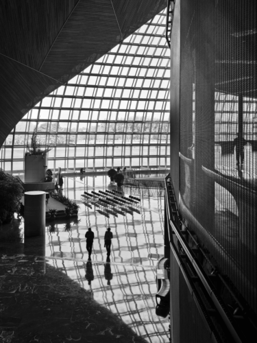 Black and white photograph of people walking through a vast modern interior with glass walls and reflections, evoking solitude, scale, and quiet observation in a personal photo essay.