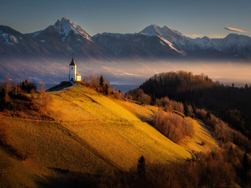 A small church perched on sunlit hills with dramatic mountains in the background, capturing timeless photography inspiration through landscape and light.