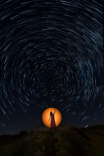 Long-exposure night photograph showing circular star trails framing a silhouetted figure illuminated by a warm glowing light, blending light painting and astrophotography, captured by Angel Torres for Questmas 2025.