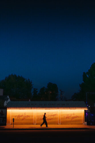 Minimalist night photograph featuring a silhouetted figure beneath a glowing bus stop against a deep blue sky, using strong negative space and contrasting color tones, captured for Questmas 2025 by William Self.