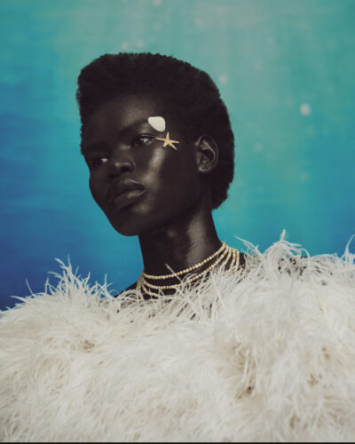 Editorial portrait of a model against a teal backdrop, styled with white feathered texture and gold accents, featuring confident gaze and refined lighting, photographed by Gustavo Chams for Questmas 2025.