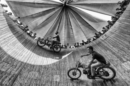Black-and-white action photograph captured inside a cylindrical wall of death arena, showing motorcyclists riding along curved vertical walls while spectators look on, creating a dramatic sense of motion and disorientation, photographed by Azim Khan Ronnie for Questmas 2025.