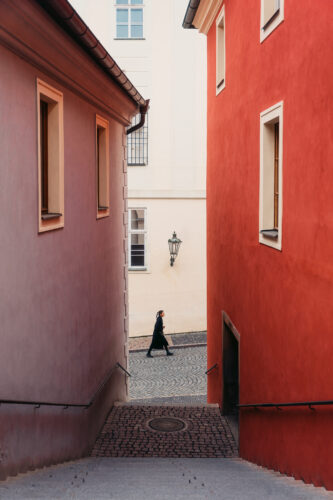 Street photography scene of a lone pedestrian walking through a narrow cobblestone alley framed by warm red and pastel buildings, using strong color contrast and natural framing, captured by Szabo Viktor for Questmas 2025.