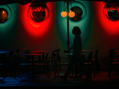 A cinematic night street scene of a person walking past café tables under red and teal neon lighting, capturing mood, color contrast, and atmosphere for a creative photo project.