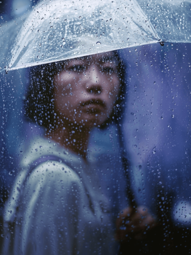A soft-focus portrait of a person holding a clear umbrella in the rain, with water droplets creating texture and emotion, illustrating atmospheric photography project ideas.