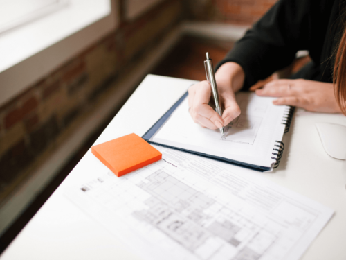 Person sketching ideas in a notebook beside printed plans and sticky notes, representing how to streamline your workflow through planning and organization.
