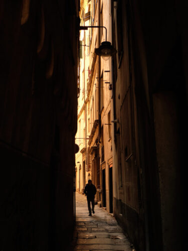 Person walking through a narrow sunlit alley, with tall buildings creating strong leading lines in composition that draw focus toward the subject.