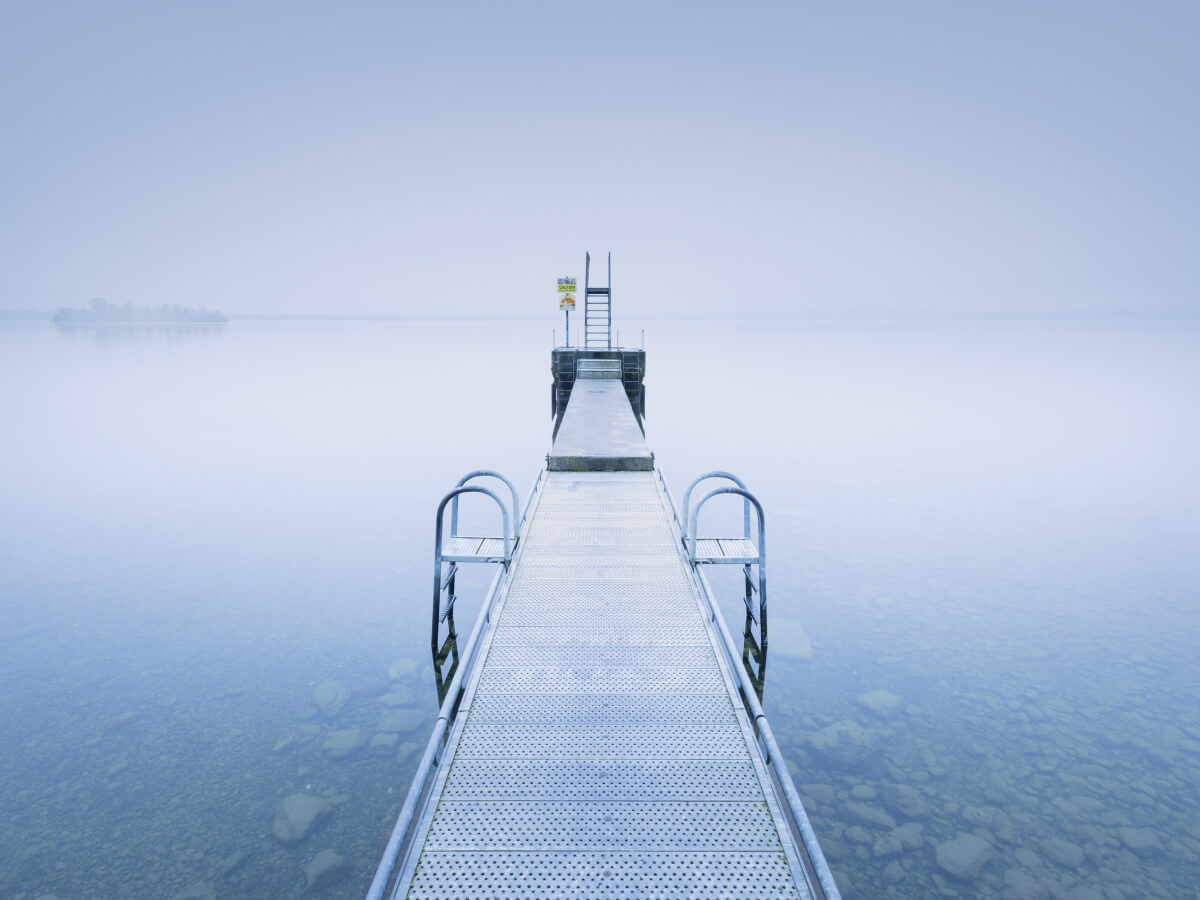 A symmetrical metal pier extending into a calm, foggy lake emphasizes repetition in photography through repeating railings and linear structure.