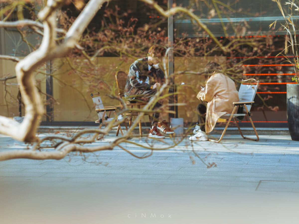 Two people sitting at an outdoor café seen through tree branches, illustrating urban candid photography in a relaxed city setting.