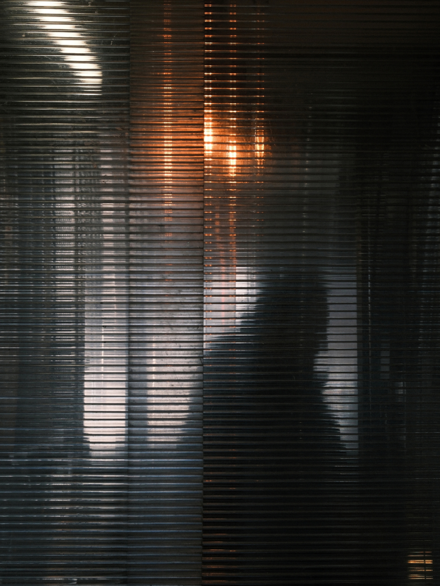 Shadow and light photography of a blurred human figure behind textured glass, with warm glowing highlights and layered reflections creating an abstract, moody atmosphere.
