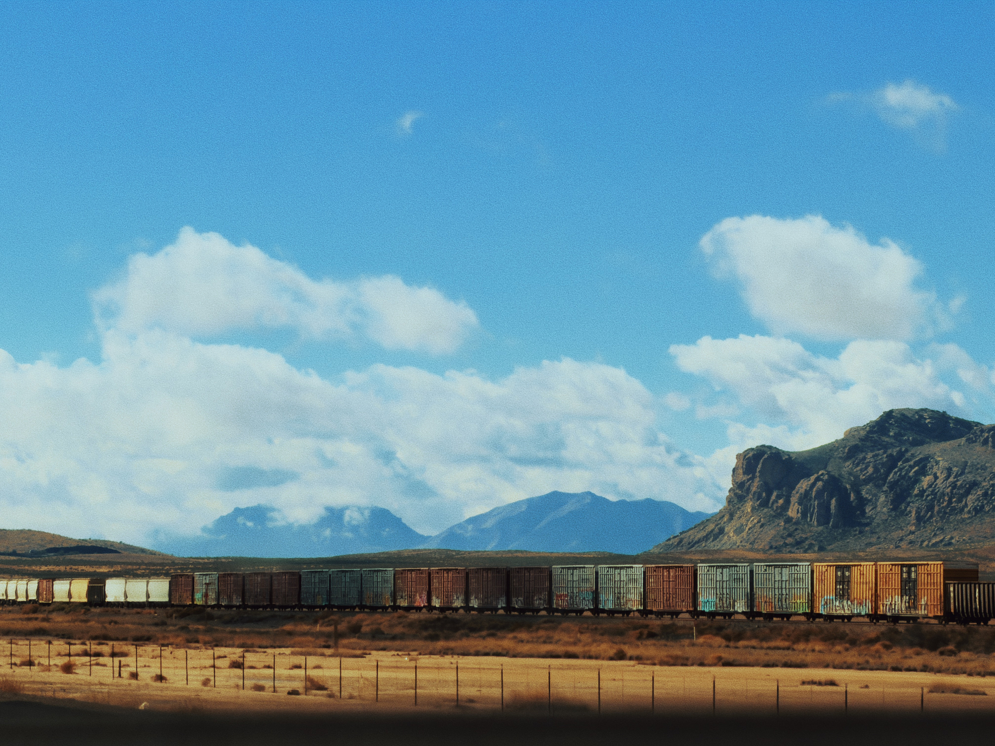 Freight train crossing a desert landscape with mountains and blue sky, captured in the cinematic photography style of Mikey Rogers.