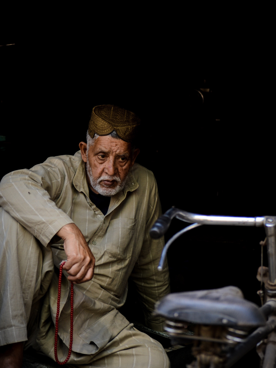 A candid portrait of an elderly man holding prayer beads, seated beside a motorcycle in dramatic shadow and light — a powerful example of candid photography capturing raw emotion and quiet contemplation.