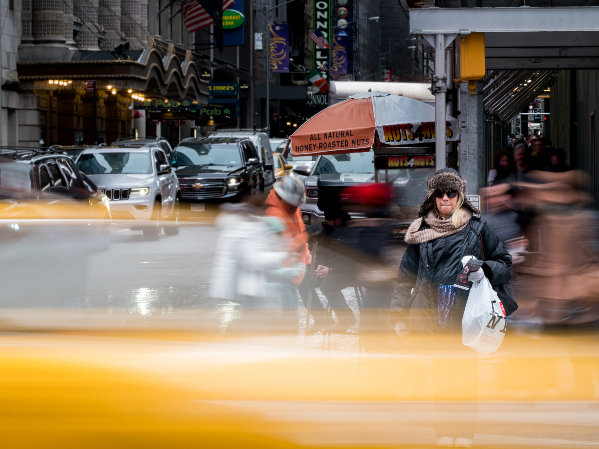 A documentary-style photographyshot of a busy New York City street, with a woman standing sharp against a blur of passing pedestrians and yellow taxis, capturing the energy and chaos of urban life.