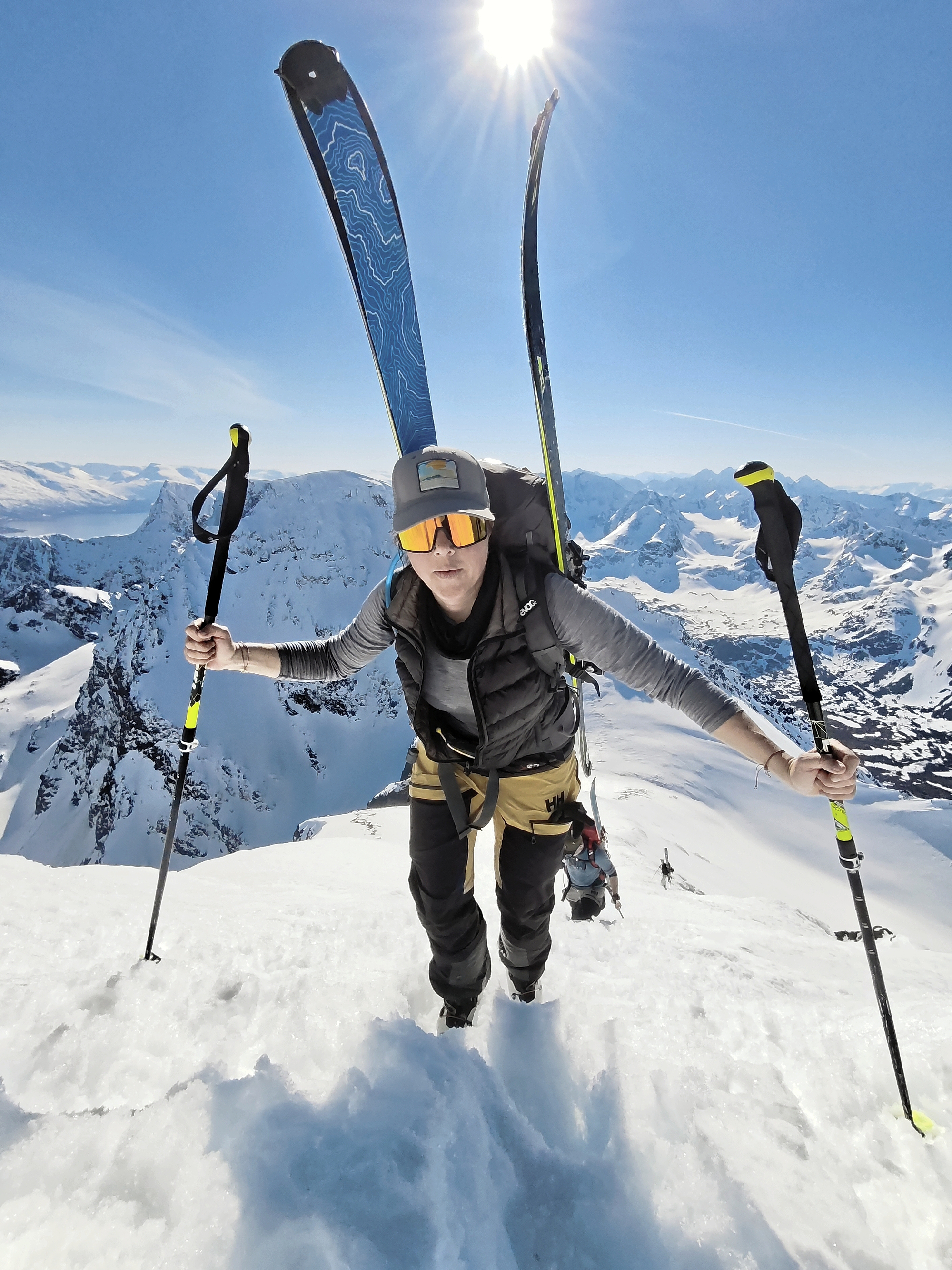A bright, front-facing close-up of a smiling mountaineer wearing a cap and sunglasses, ascending a snowy incline.